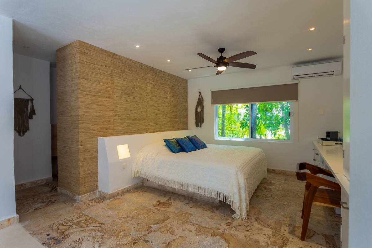 Une chambre à coucher avec un lit, un ventilateur de plafond, une fenêtre donnant sur un paysage verdoyant et un sol en pierre.
