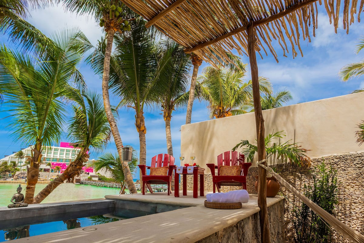 Un espace aménagé au bord de la piscine, avec des chaises rouges, une petite table sur laquelle se trouvent une bouteille et un verre, et un toit de chaume.
