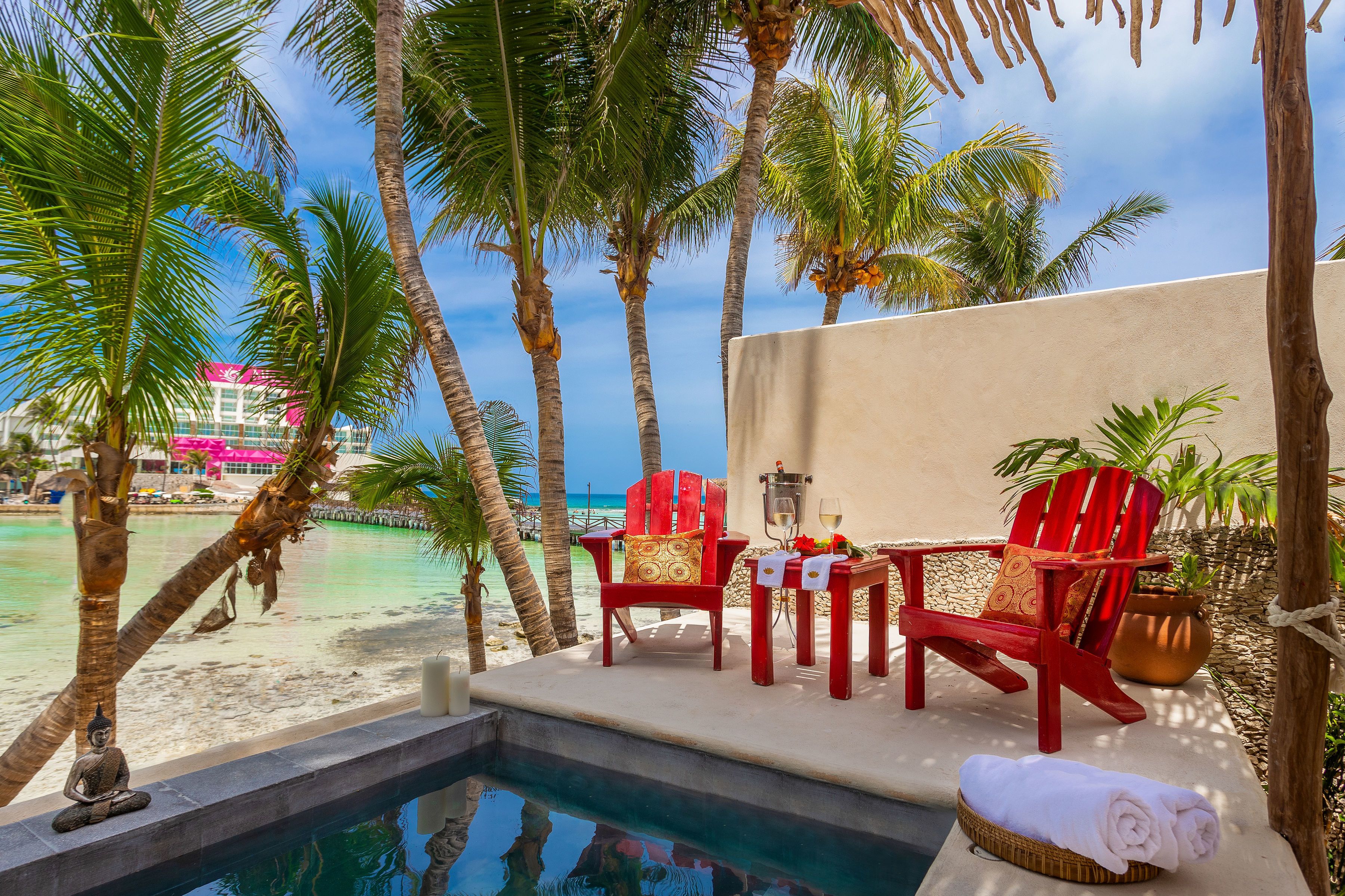 A poolside area with a view of the beach, red chairs, a small table with a candle, and a statue.