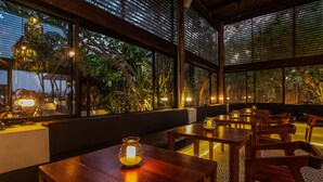 Un restaurant avec des tables et des chaises en bois, de grandes fenêtres équipées de stores et une vue sur la verdure environnante.