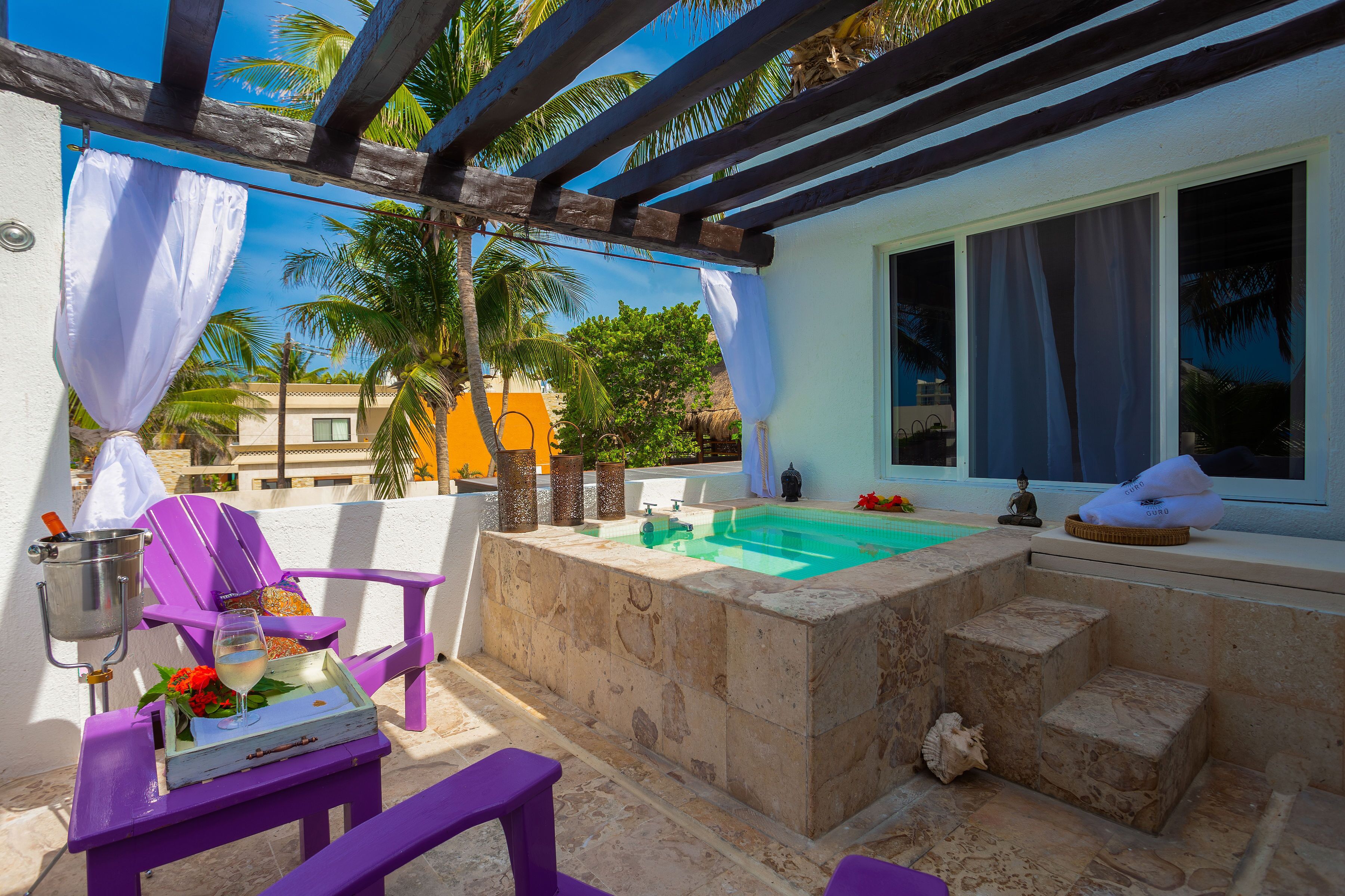 Un área de piscina privada con un jacuzzi de piedra, una silla lounge morada y una mesa con una bebida y flores.