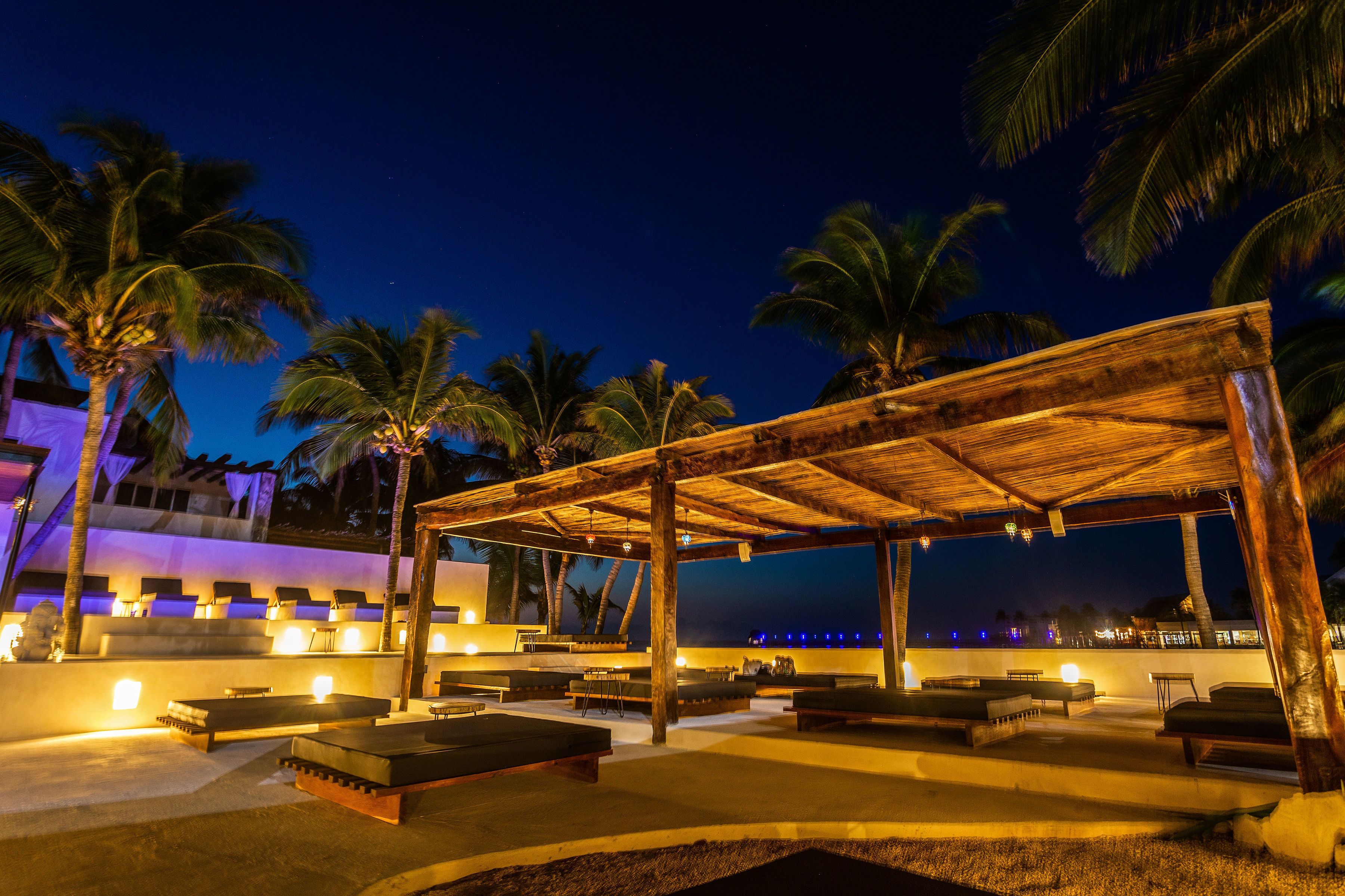 A covered outdoor seating area with lounge chairs and a wooden pergola, surrounded by palm trees and a pool.