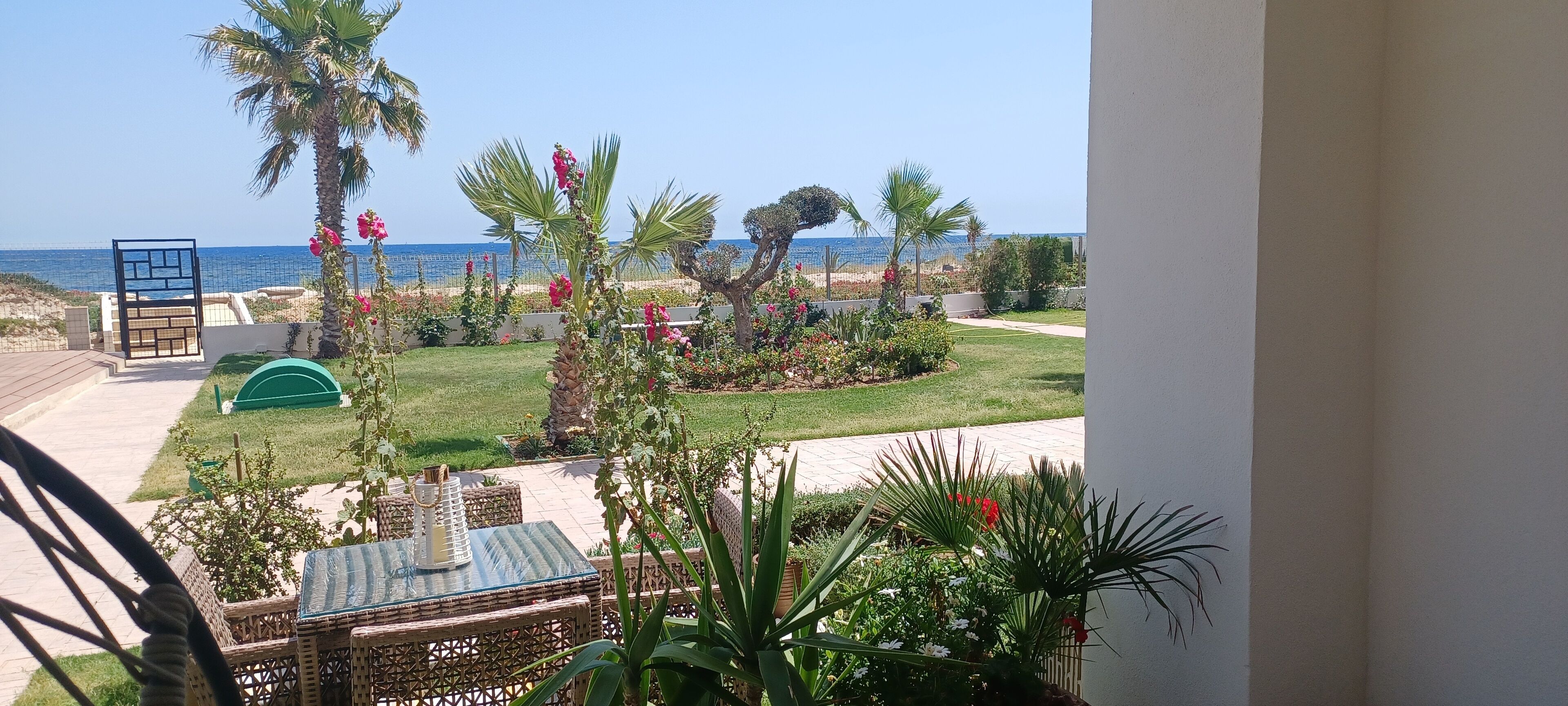 A balcony with a view of the sea, a garden with palm trees, and a patio area with wicker furniture.