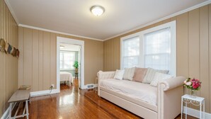A room with a beige sofa, wooden flooring, a white side table with a vase of flowers, and a door leading to another room. - Insta Worthy Stay! - Fun in Downtown Raleigh! (Raleigh)