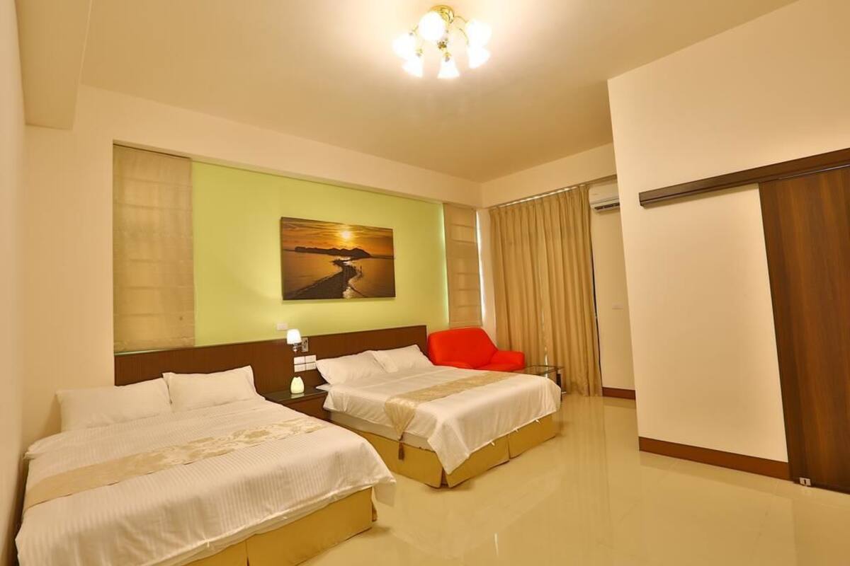 A hotel room with two beds, a red chair, a painting on the wall, and a large window with curtains.