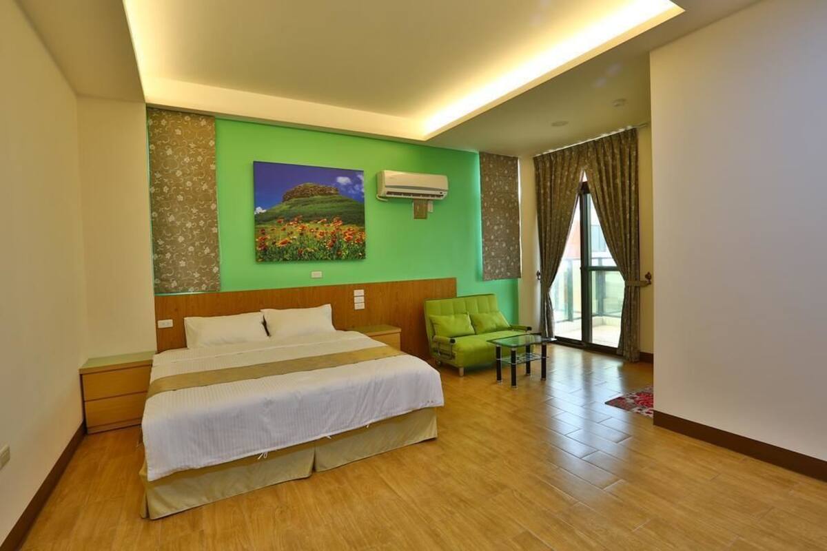 A hotel room with a large bed, a green sofa, a small table, and a painting on the wall.