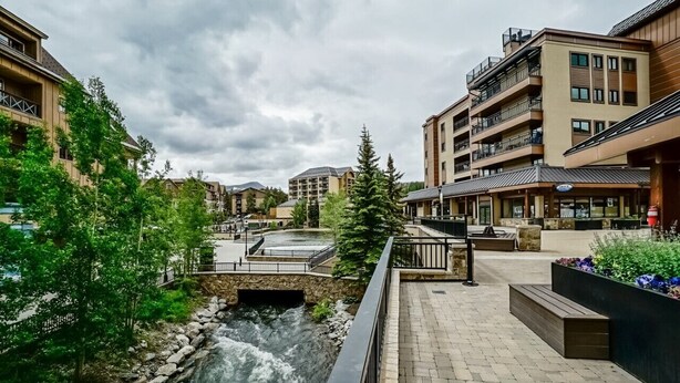 A riverside walkway with stone steps, a small bridge, and buildings with balconies and outdoor seating areas. - Luxury|In Town|Peak 9|Ski-In/Out (Breckenridge)