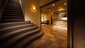 A staircase leading to a lobby with a checkered floor, a stone wall, and a seating area with a fireplace. - Hoozue No Toki (Izu)
