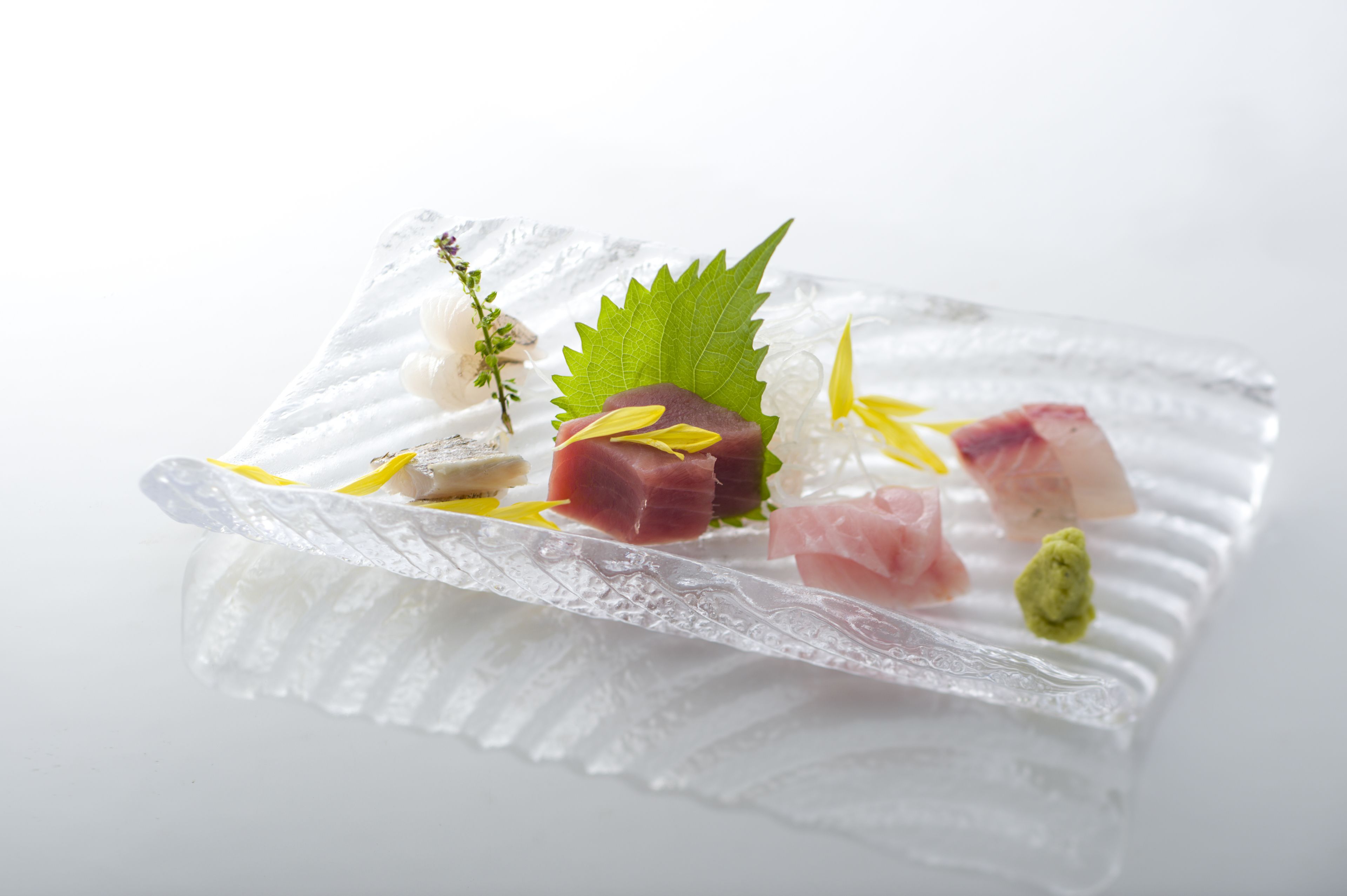 A clear glass plate with various food items, including fish and garnishes, on a white background.