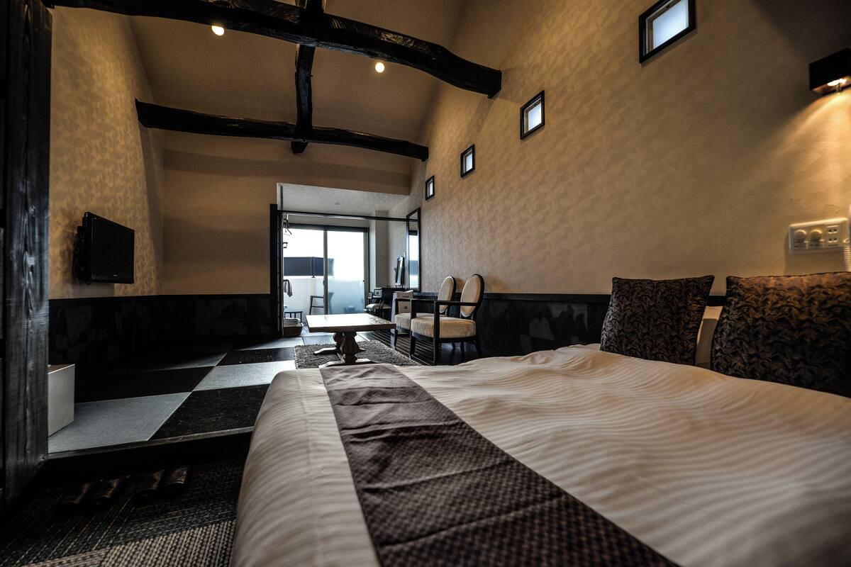 A hotel room with a large bed, a desk, and a chair. There is a window with a view of the outside.