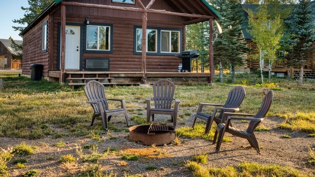 A wooden cabin with a porch, three Adirondack chairs, and a fire pit.