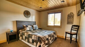 A bedroom with a wooden ceiling, a bed with a plaid bedspread, a nightstand with a tissue box, a window with blinds, and a chair.