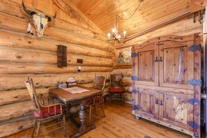 Interior rústico de una cabaña de troncos con una mesa de comedor de madera, sillas, una araña y un gran armario de madera.
