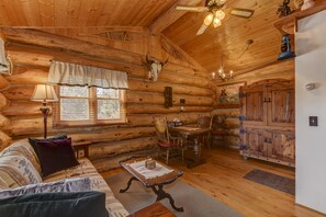 Interior de una cabaña de troncos con techo de madera, una mesa de comedor con sillas, un sofá con cojines y un armario.