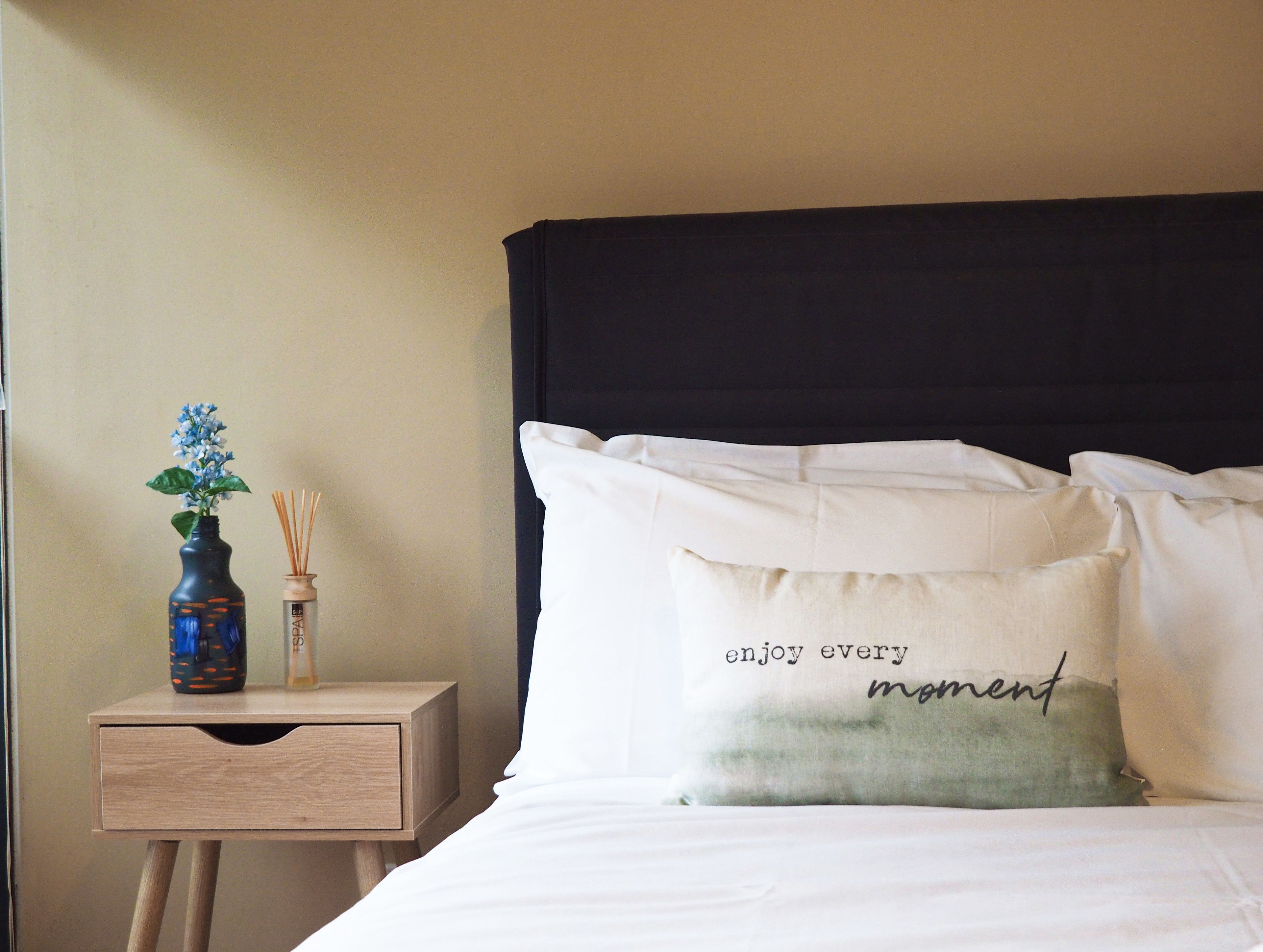Una cama bien hecha con almohadas blancas y verdes, una mesita de noche de madera con un florero y difusor de aroma, y un cabecero oscuro.