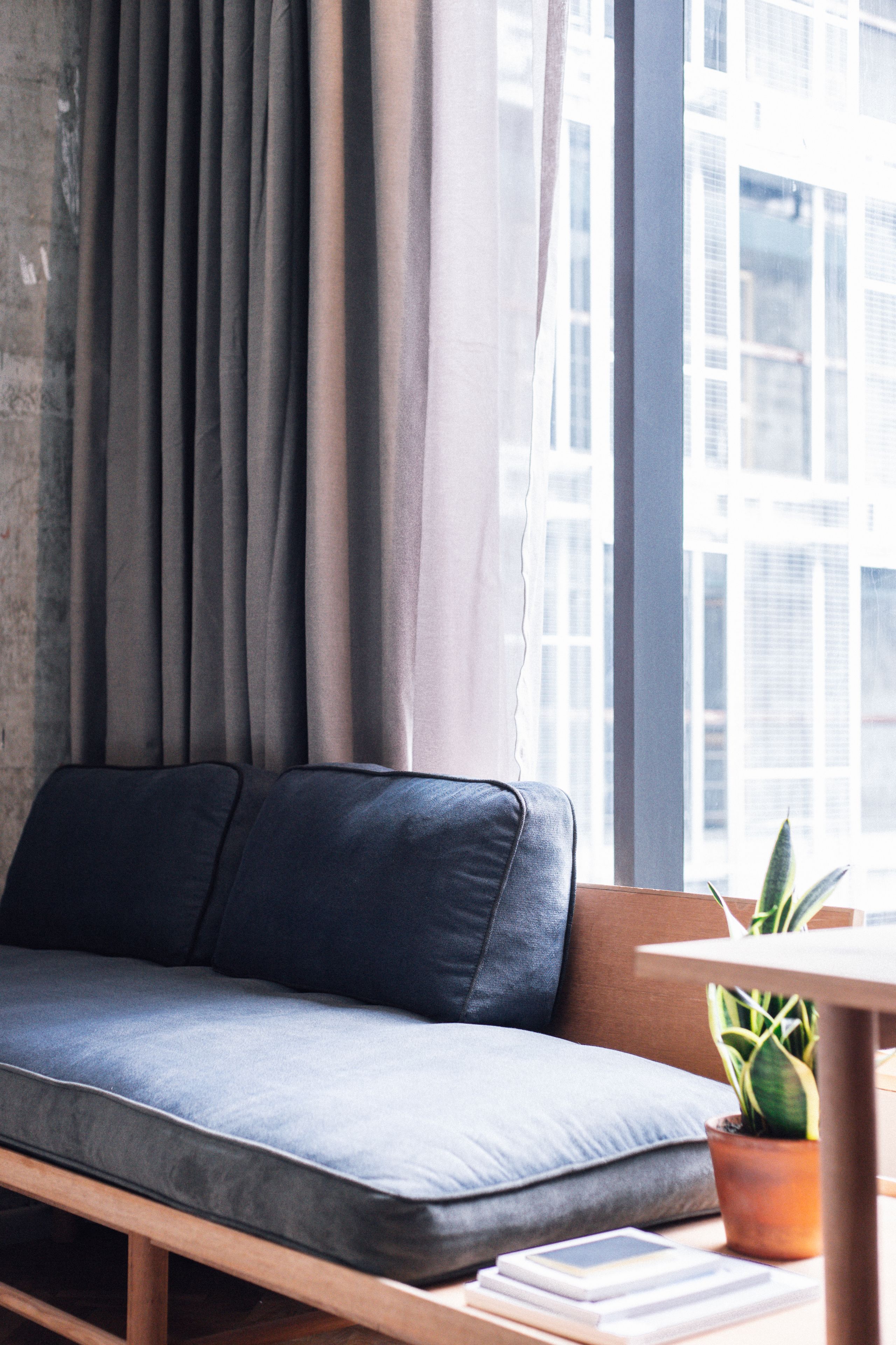 A modern living room with a grey sofa, a small table with a plant, and a view of buildings through large windows.