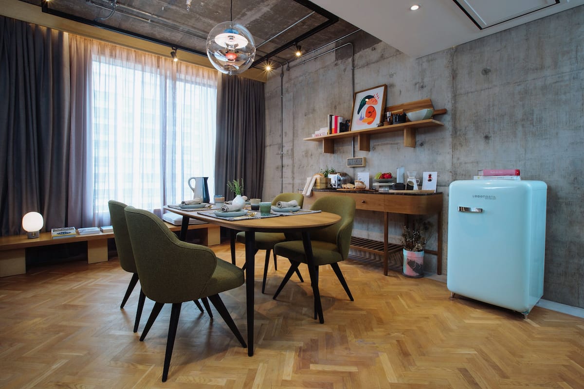 A modern dining area with a wooden table, green upholstered chairs, a light blue refrigerator, and a concrete wall with a shelf holding books and decorative items.