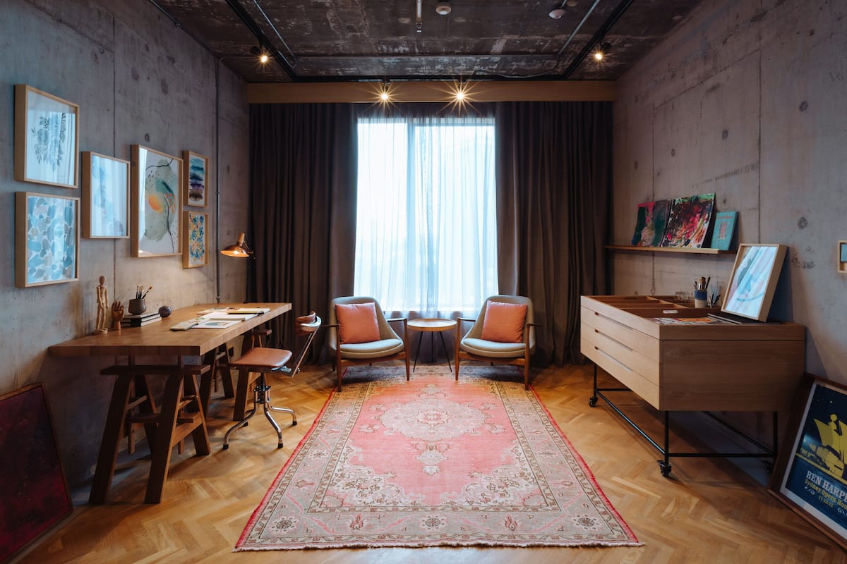 A room with a wooden desk, two chairs, a small table, a large window with curtains, and artwork on the walls.