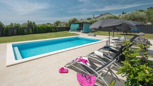Una piscina rectangular con sillones de descanso y sombrillas, rodeada de una valla y vegetación.