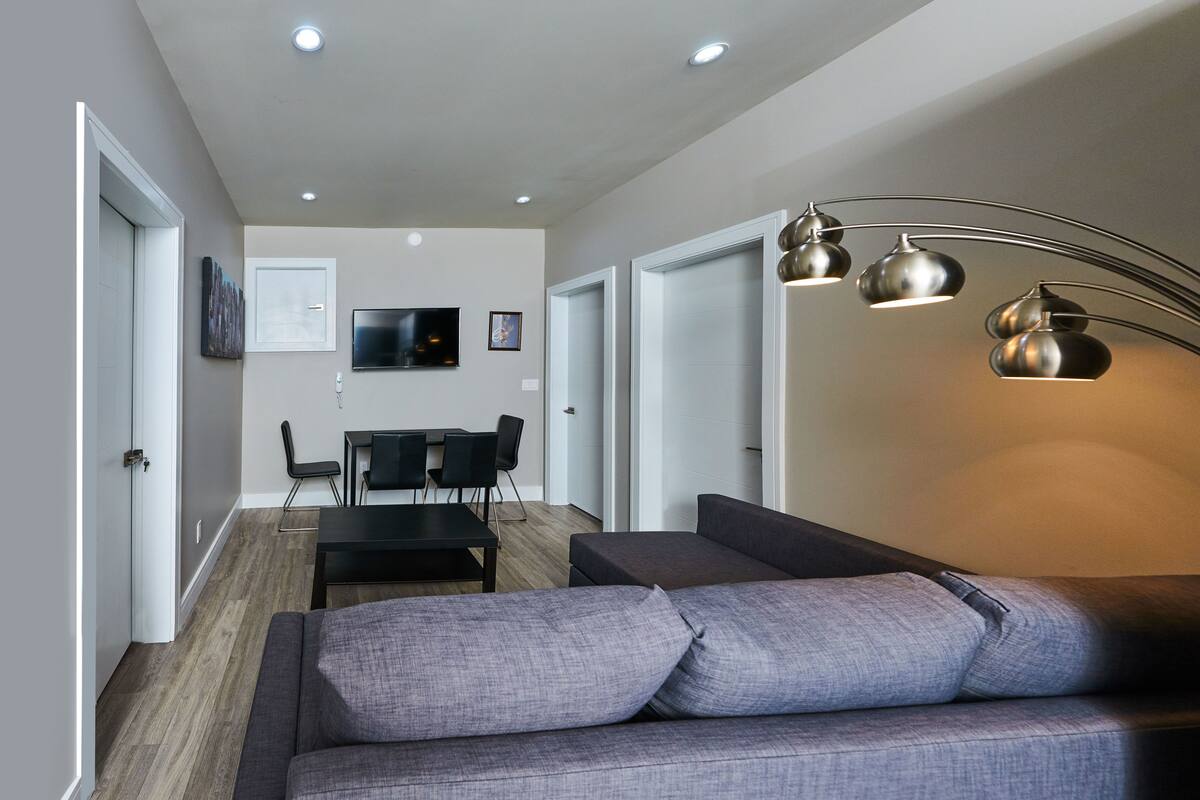 A modern living room with a grey sofa, a dining area with a black table and chairs, and recessed ceiling lighting.