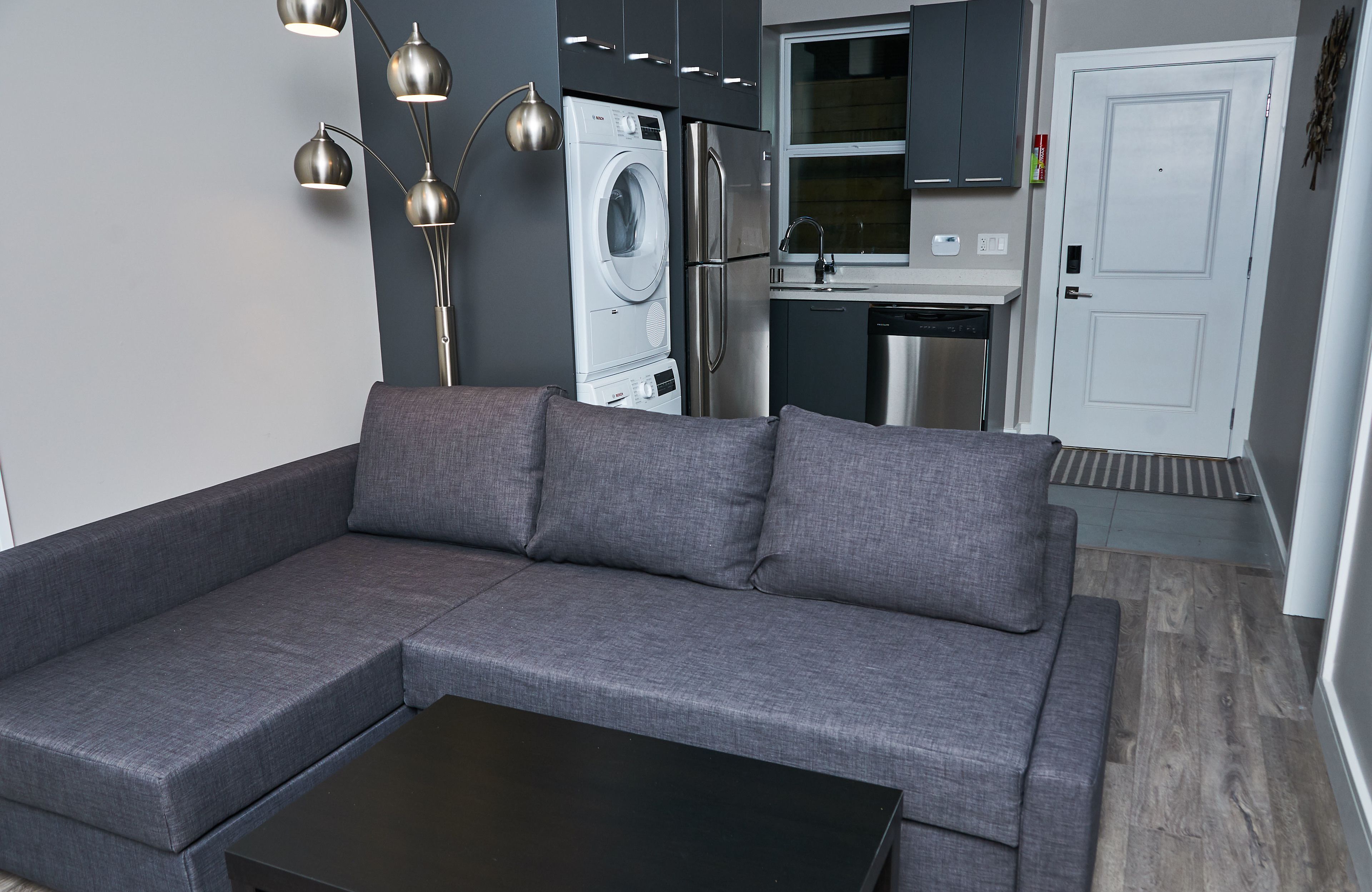 Un salon moderne avec un canapé d’angle gris, une table basse noire, un lave-linge et un réfrigérateur.