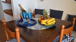 Et træspisebord med en blå bakke, to blå glas, en champagneflaske og en kurv med frugt, herunder bananer, appelsiner og en ananas.