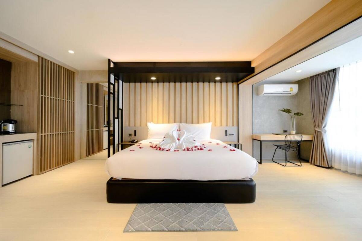 A modern hotel room with a large bed, a small desk, a kitchenette, and a view of the city.