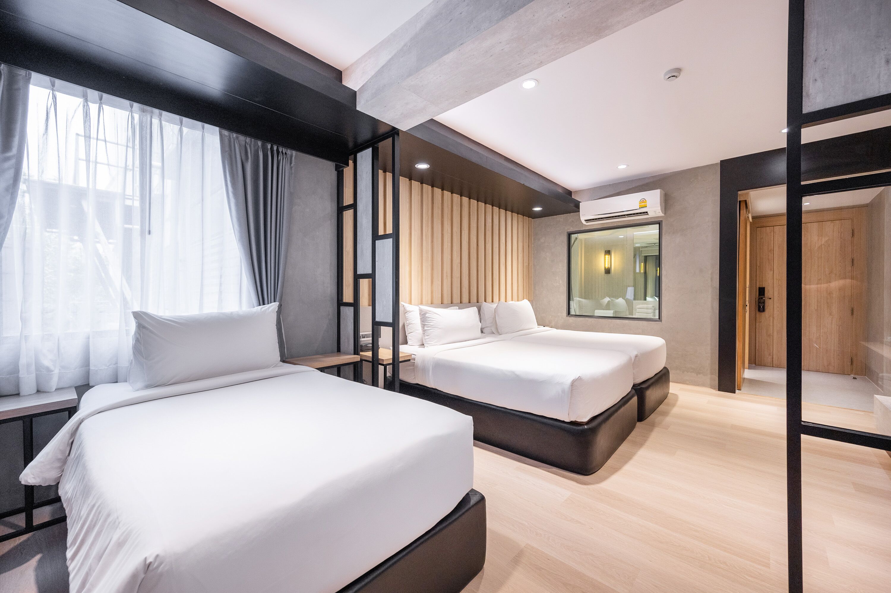 Une chambre d’hôtel moderne avec deux lits, une grande fenêtre avec des rideaux, un mur revêtu de panneaux en bois et un miroir au-dessus des lits.