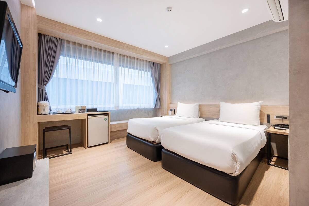 A modern hotel room with two beds, a desk with a computer, a small refrigerator, and a TV mounted on the wall.