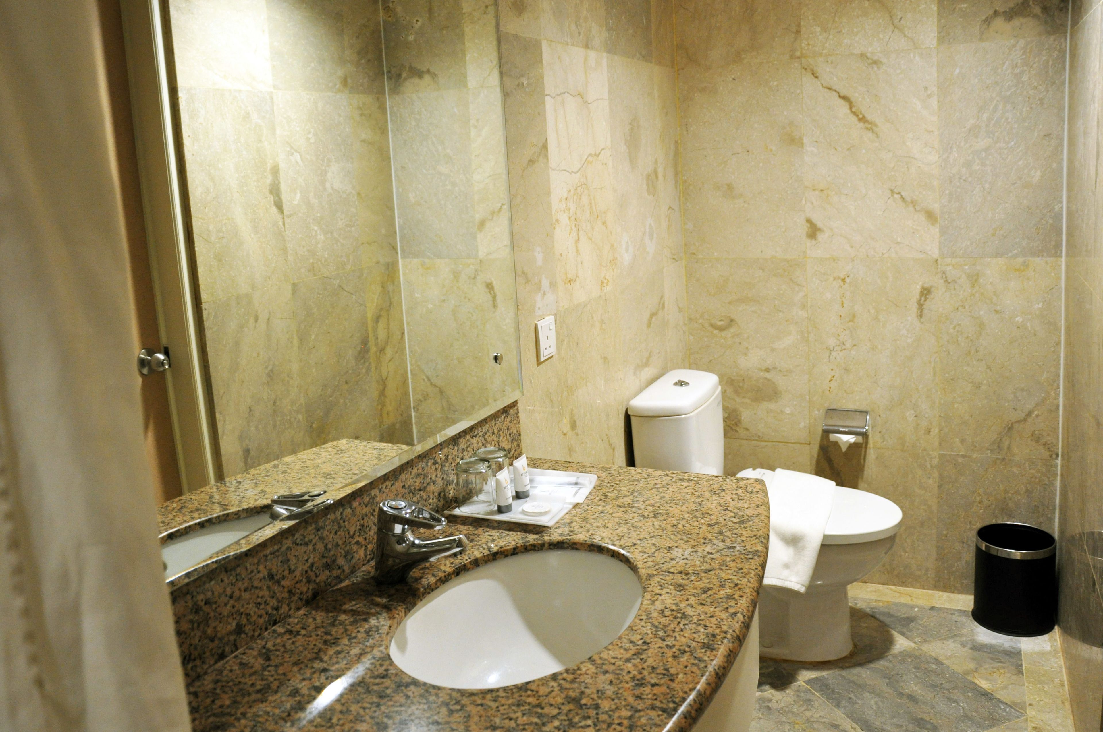 A bathroom with a granite countertop sink, a mirror, a toilet, and a trash can.