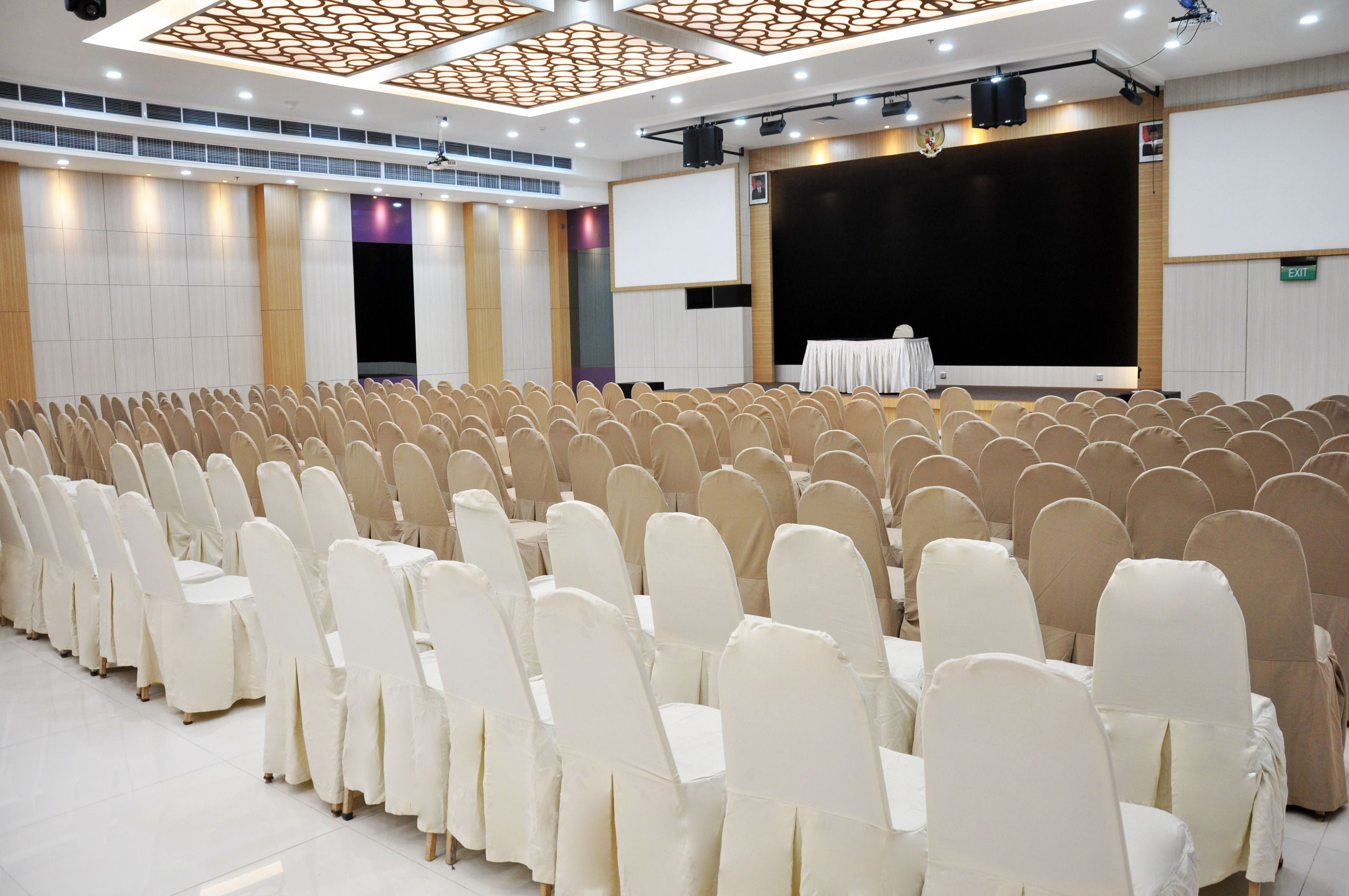 Sala de conferências com fileiras de cadeiras, palco com projetor e uma tela grande.