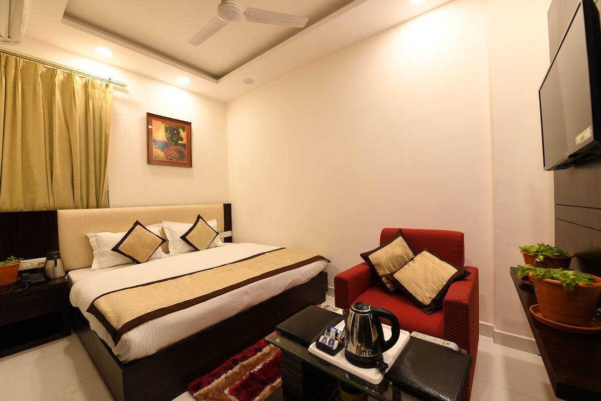 A hotel room with a bed, a red armchair, a coffee table, a TV, and a painting on the wall.