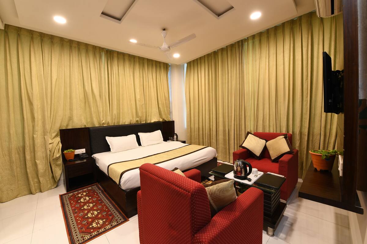 A hotel room with a large bed, a red armchair, a small table with a coffee pot, and curtains.