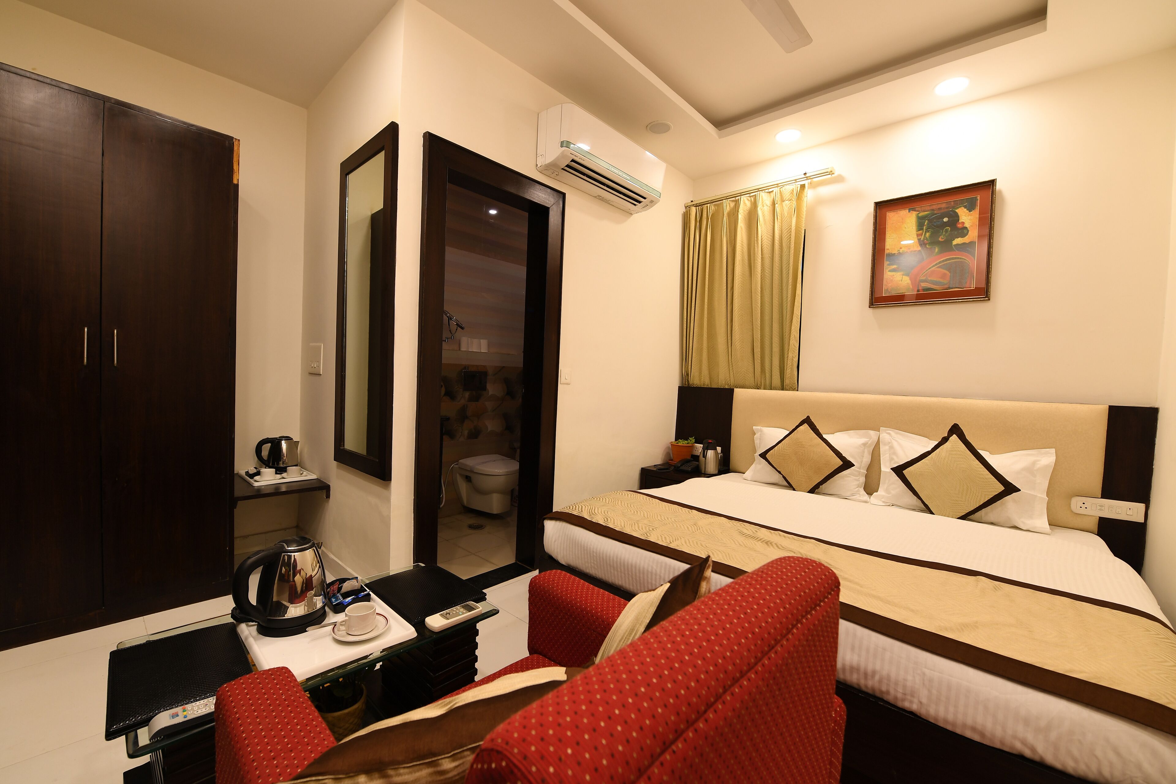 A hotel room with a bed, a desk with a kettle, a chair, a wardrobe, and an air conditioning unit.