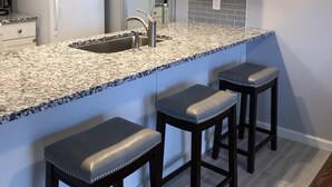 Een moderne keuken met granieten aanrechtbladen, witte kasten en een roestvrijstalen spoelbak. Drie barkrukken met beklede zittingen.