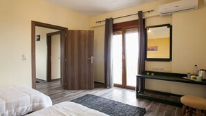 Une chambre d’hôtel avec une porte en bois, une porte-fenêtre coulissante, un lit, un petit tabouret, une étagère avec des articles de toilette et un miroir.
