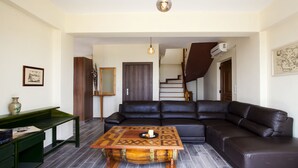 Un salon avec un canapé en cuir noir, une table basse en bois, un banc vert et un escalier menant à l’étage supérieur.