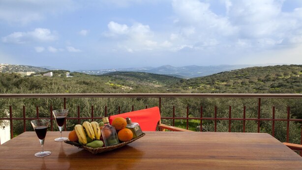 Une table en bois, sur laquelle se trouve un panier de fruits, deux verres à vin et un coussin rouge, est placée sur un balcon donnant sur un paysage montagneux.