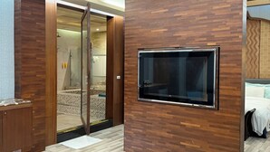 Une chambre d’hôtel moderne avec un plancher en bois, un téléviseur fixé sur un panneau en bois, une table en bois avec une boîte de mouchoirs et une cabine de douche en verre.