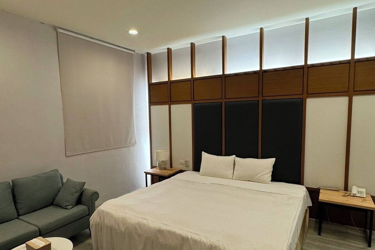 Habitación de hotel con una cama grande, un sofá, una mesita redonda y una mesita de noche con lámpara y teléfono.
