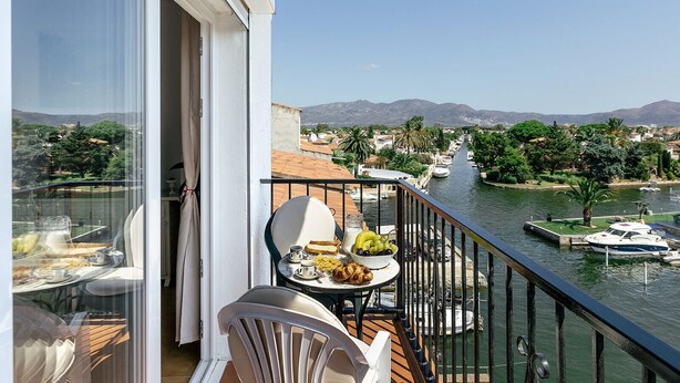 Un balcon avec une table dressée pour le petit-déjeuner, donnant sur un canal où naviguent des bateaux, et avec une vue sur la ville en arrière-plan.