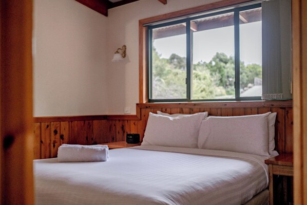 Ein Schlafzimmer mit Holzkopfteil, einem großen Fenster mit Blick ins Grüne und einem ordentlich bezogenen Bett mit einem weißen Kopfkissen und einem gefalteten weißen Handtuch.