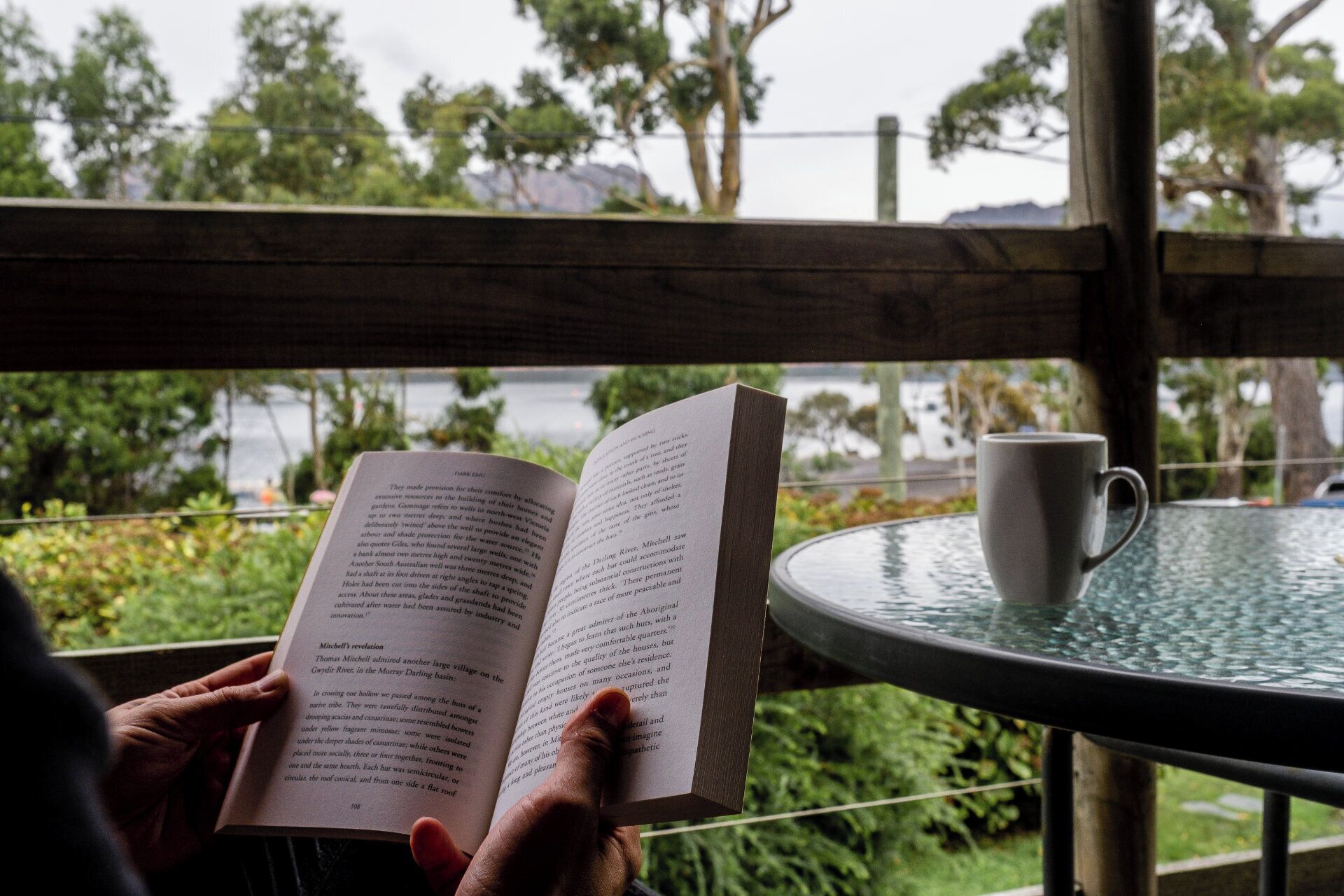 A person reading a book at a table with a cup, outdoors with greenery and a view of a body of water.