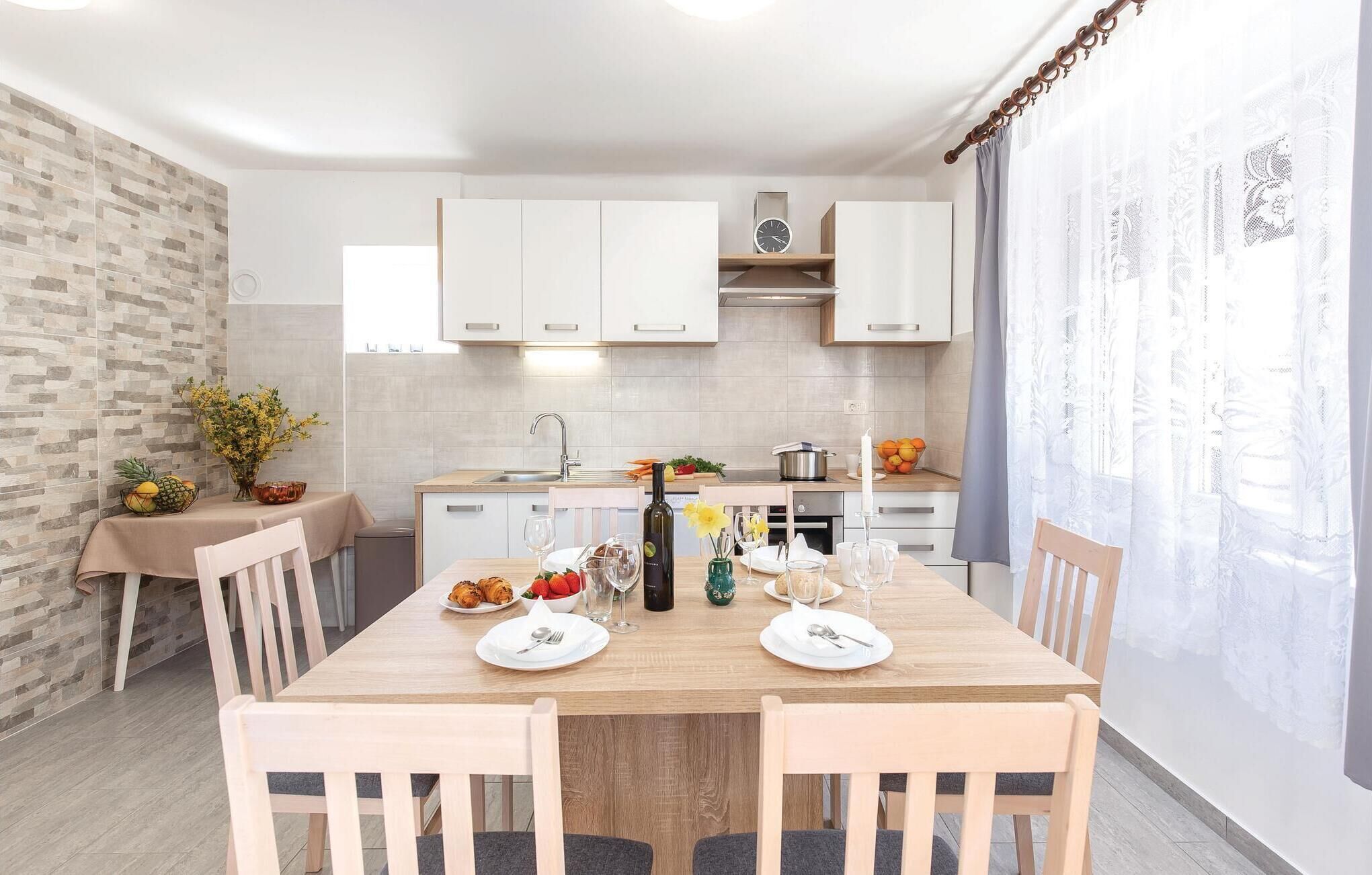 A modern kitchen with white cabinets, a wooden dining table, and a view of the outdoors through sheer curtains.