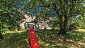 A playground with a red slide and a green swing set, surrounded by trees and a house in the background. - 2 bedroom pet friendly home in Skrljevo (Skrljevo)