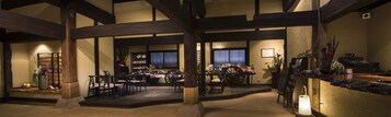 Un interior espacioso con techos altos y vigas de madera a la vista, que cuenta con una zona de estar central con mesas y sillas, rodeada de estanterías y objetos decorativos.