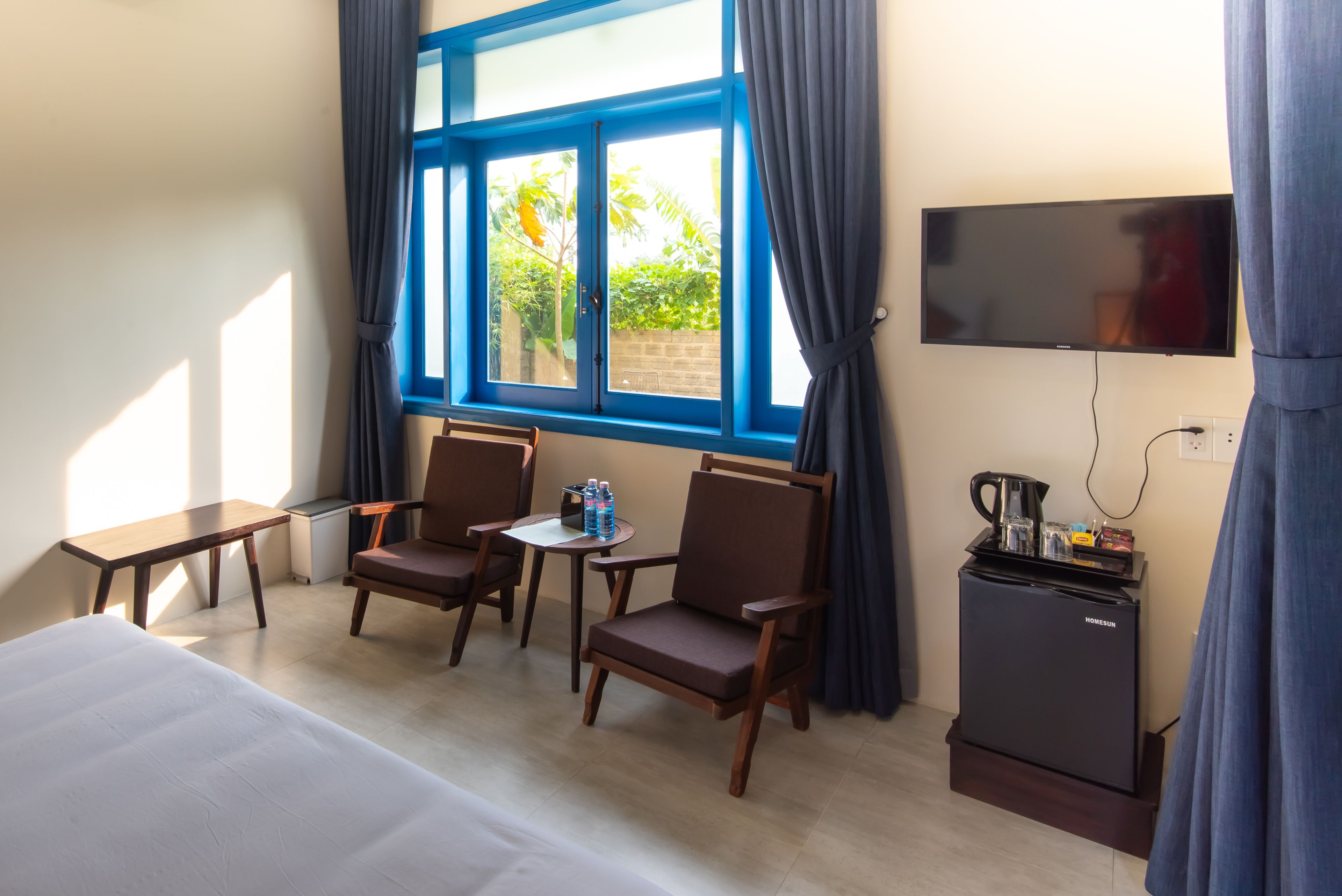 A hotel room with a bed, two chairs, a small table, a TV, and a minibar.