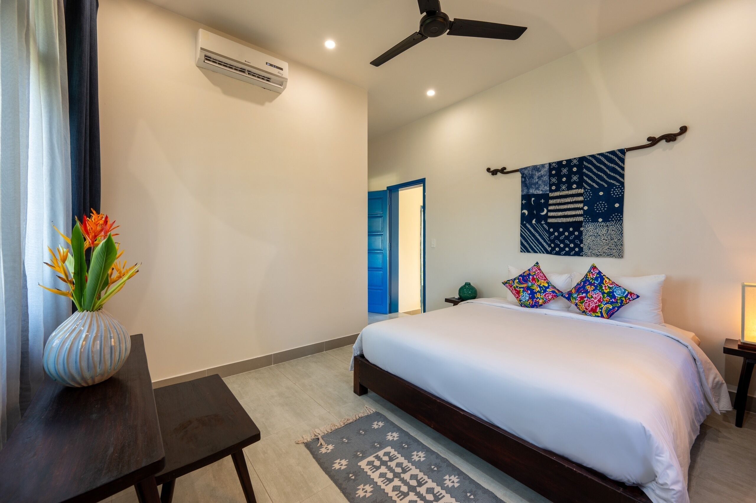Un dormitorio con cama, ventilador de techo, aire acondicionado, un jarrón con flores, una puerta azul y un tapiz en la pared.