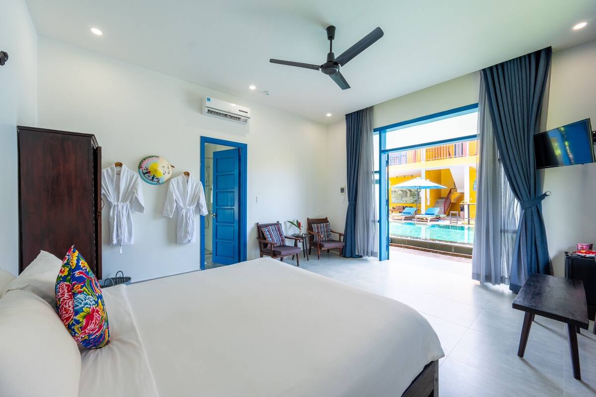 A hotel room with a large bed, a ceiling fan, an air conditioning unit, a TV, and a view of a pool area.