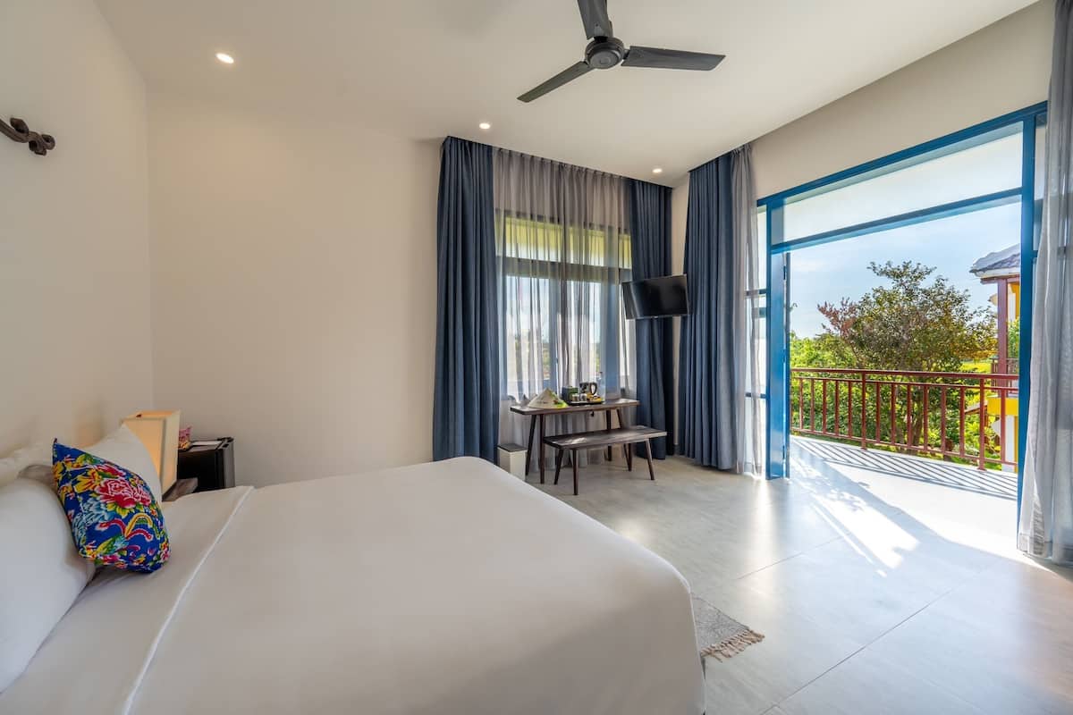 A hotel room with a bed, a desk, a chair, a TV, and a balcony with a railing.